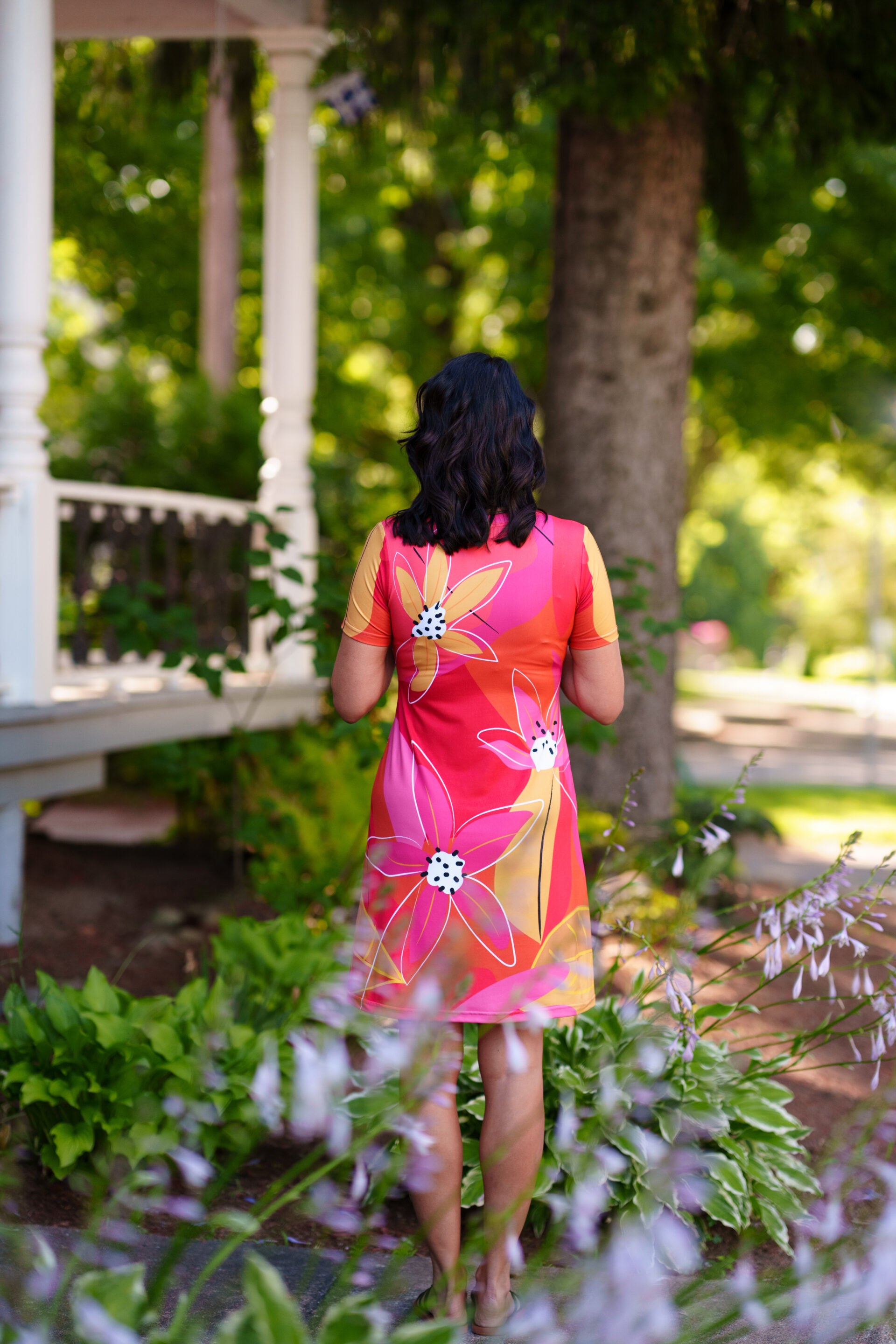 Back view of a woman wearing the Aperol Dress from Rien ne se Perd, a short flared dress with a V-neck, short sleeves, and a colourful Pink floral pattern She is standing in a garden setting.