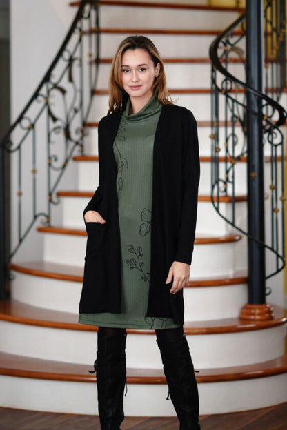 A woman wearing the Aurele Dress by Rien ne se Perd in Khaki under a black cardigan,  standing in front of a staircase. 