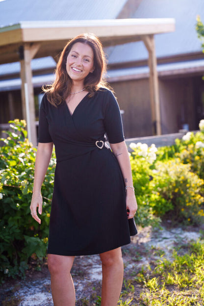 A woman wearing the Bachata Dress by Rien ne se Perd, a Black wrap dress with a V-neck, waist tie with a heart-shaped coconut shell buckle, and short A-line skirt. She is standing outside. 