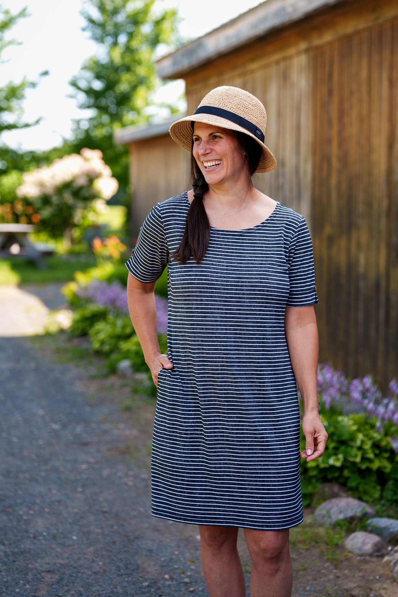 
A woman wearing the Boogie Dress by Rien ne se Perd in Grey/White Stripe, a t-shirt dress with a wide neck, side pocket, and slit and coconut button detail on the short sleeves. She is standing outside in front of a wooden building. 