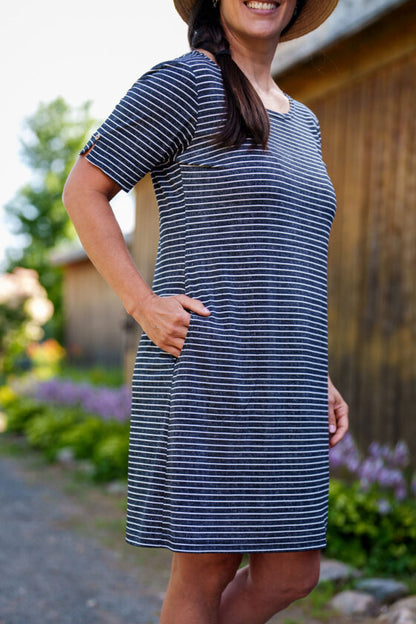 3/4 view of a woman wearing the Boogie Dress by Rien ne se Perd in Grey/White Stripe, a t-shirt dress with a wide neck, side pocket, and slit and coconut button detail on the short sleeves. She is standing outside. 