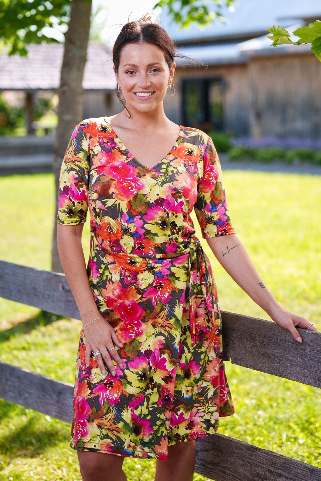 A woman wearing the Cha Cha Cha Dress by Rien ne se Perd, a faux-wrap dress in a floral pattern with a V-neck, tie at the waist, and knee-length A-line skirt. She is standing by a fence.