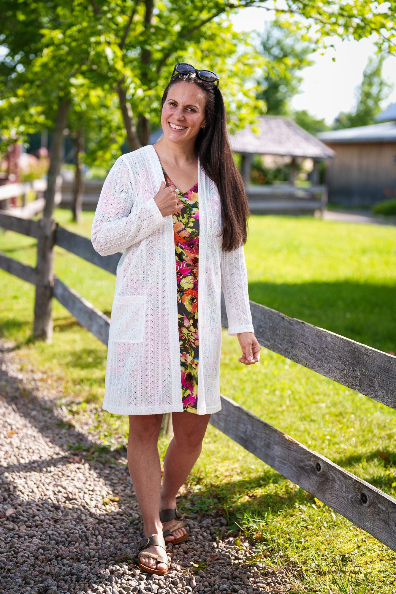 A woman wearing the Cha Cha Cha Dress by Rien ne se Perd, a faux-wrap dress in a floral pattern with a V-neck, tie at the waist, and knee-length A-line skirt. She wearing it with a white sweater and is standing by a fence.