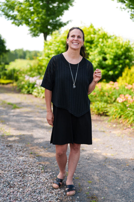 A woman wearing the Menuet Bolero- Poncho by Rien ne se Perd, a Black woven lightweight poncho. She's wearing it over a black dress and standing outside. 