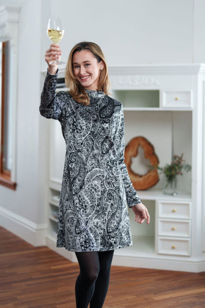 A woman wearing the Frida Dress by Rien ne Perd in Black and White Velvet stands in front of a white cabinet, holding a glass of wine.