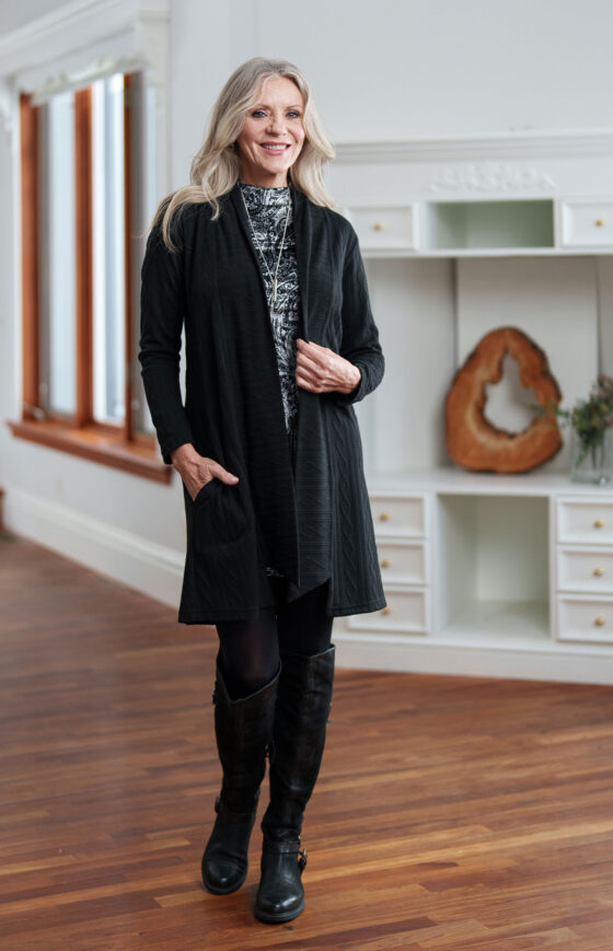 A woman wearing the Frida Dress by Rien ne Perd in Black and White Velvet under a Black cardigan stands in front of a white cabinet  