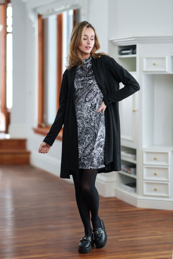 A woman wearing the Frida Dress by Rien ne Perd in Black and White Velvet under a Black cardigan stands in front of a white cabinet  