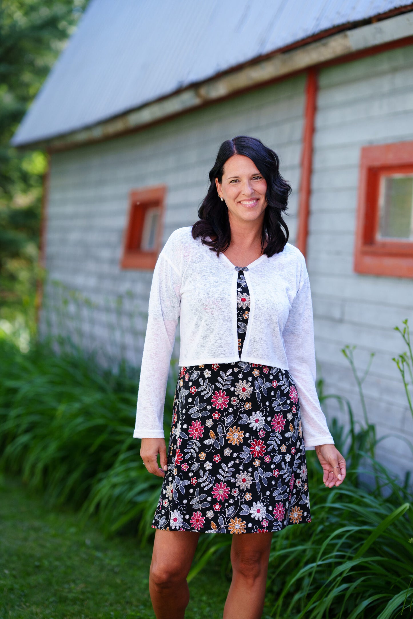 A woman wearing the Grenadine Dress by Rien ne se Perd in Floral print,  a short A-line dress with bell sleeves and a scoop neck. She is wearing it under a white cardigan and  standing in front of a rustic building with red windows.