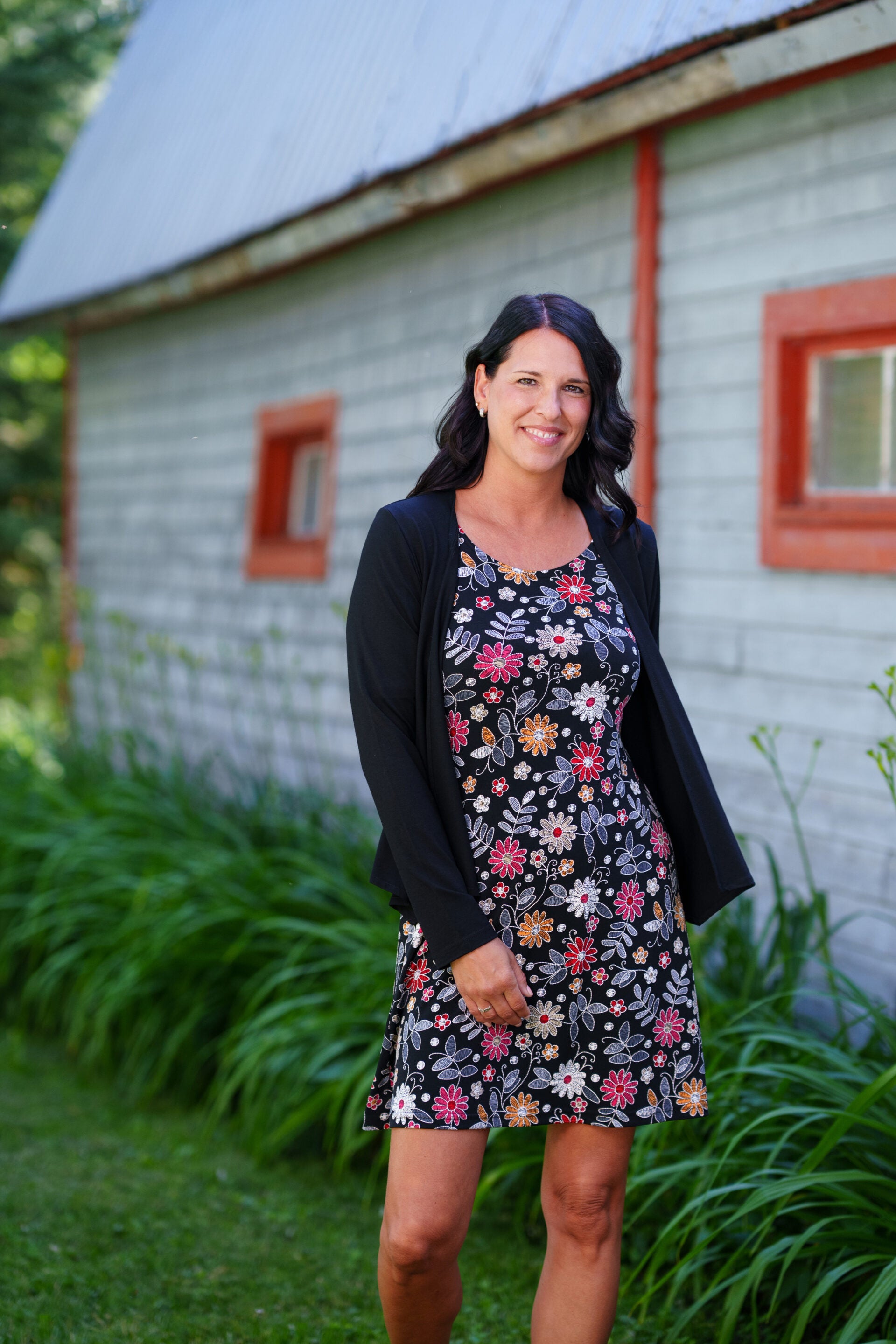 A woman wearing the Grenadine Dress by Rien ne se Perd in Floral print,  a short A-line dress with bell sleeves and a scoop neck. She is  wearing it under a black cardigan and standing in front of a rustic building with red windows.