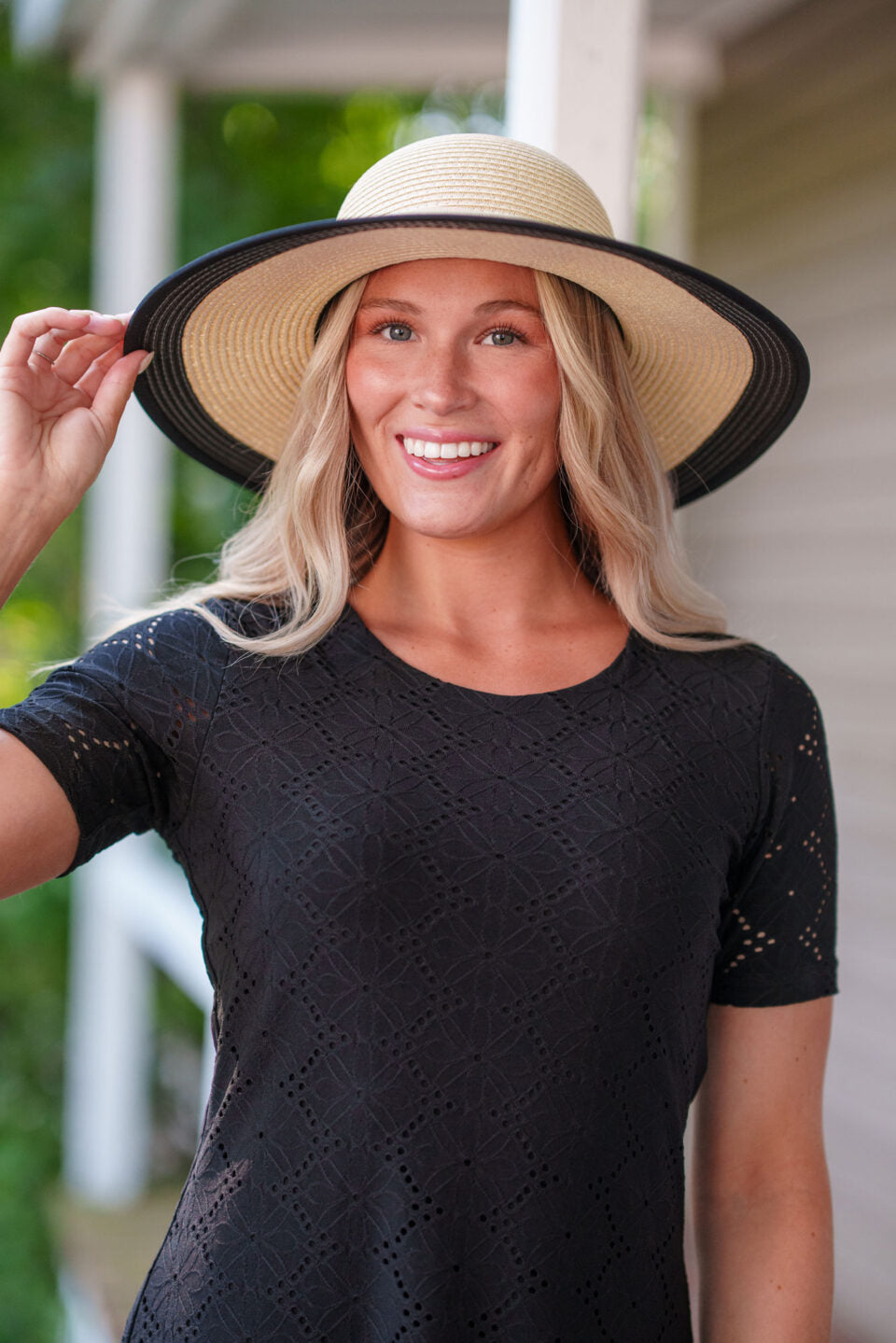 Close up view of a woman wearing the Litchi Dress by Rien en se Perd in Black, a semi-fitted short dress with short sleeves and an openwork daisy fabric. She is wearing it a hat and standing on a porch.