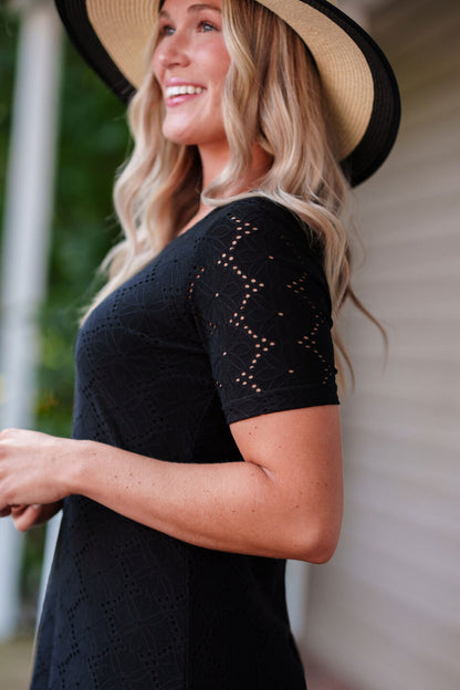Close up side view of a woman wearing the Litchi Dress by Rien en se Perd in Black, a semi-fitted short dress with short sleeves and an openwork daisy fabric. She is wearing it a hat and standing on a porch.