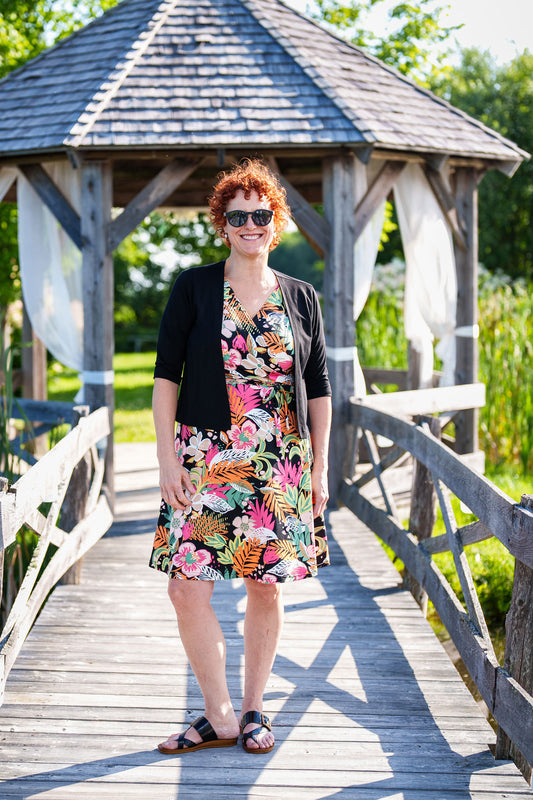 A woman wearing the Salvador Bolero in Black from Rien ne se Perd, featuring 3/4 sleeves and an angled hem, over the Macarena Dress. She is standing on a dock in front of a wooden gazebo. 