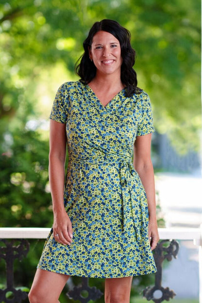 A woman wearing the Margarita Dress by Rien ne se Perd in Blue/Green floral print, with a faux wrap neckline, short sleeves, and short a-line skirt. She is standing on a porch. 
