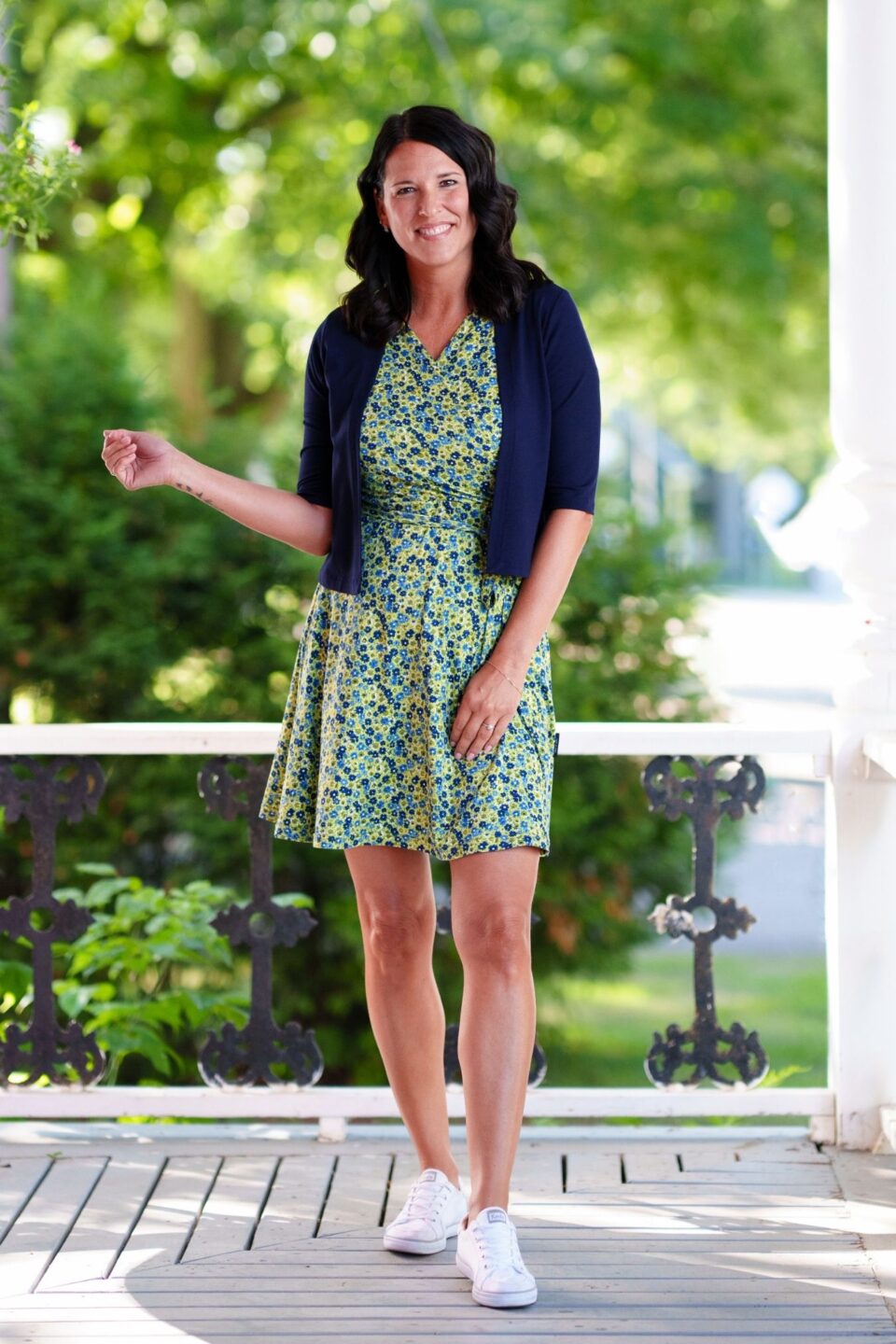 A woman wearing the Margarita Dress by Rien ne se Perd in Blue/Green floral print, with a faux wrap neckline, short sleeves, and short a-line skirt. She is wearing it with a navy bolero and standing on a porch. 
