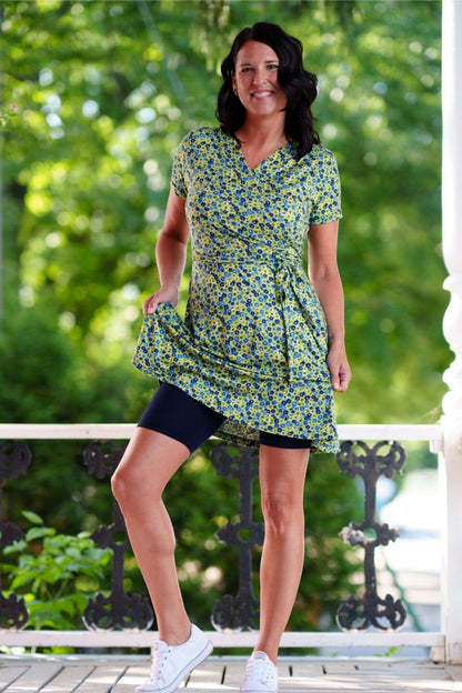 A woman wearing the Margarita Dress by Rien ne se Perd in Blue/Green floral print, with a faux wrap neckline, short sleeves, and short a-line skirt. She is wearing it with black bike shorts and standing on a porch. 