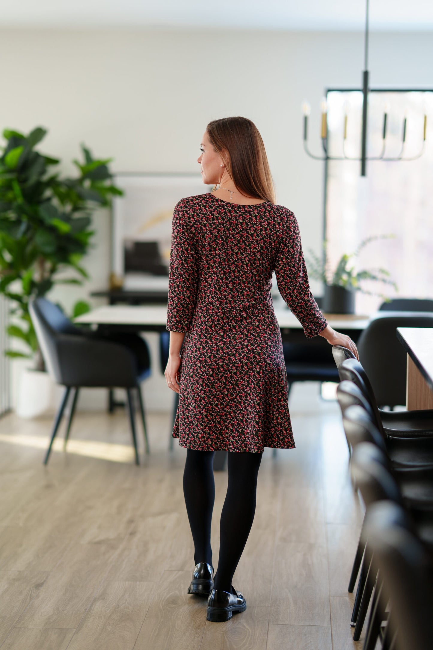 Back view of a woman wearing the Mattawin Dress from Rien ne se Perd in Red Floral, an above-the-knee length faux-wrap dress with three-quarter sleeves. She is standing in a modern, softly blurred dining room setting.
