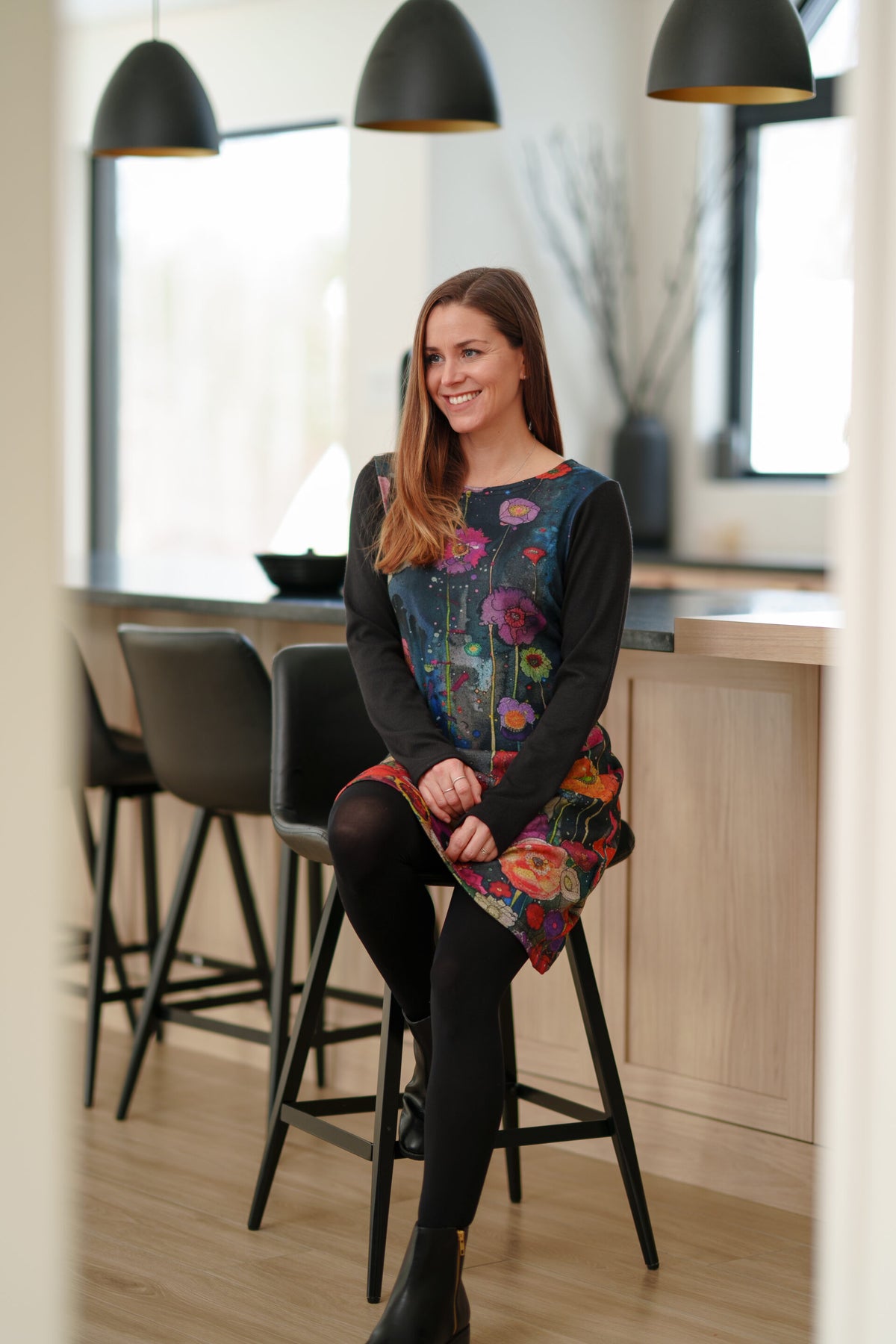 A woman wearing the Mistassini Dress by Rien ne se Perd in Floral Watercolour, an A-line knee-length dress with a vibrant floral print on the main body and contrasting black sleeves. She sitting by a counter in a kitchen setting. 