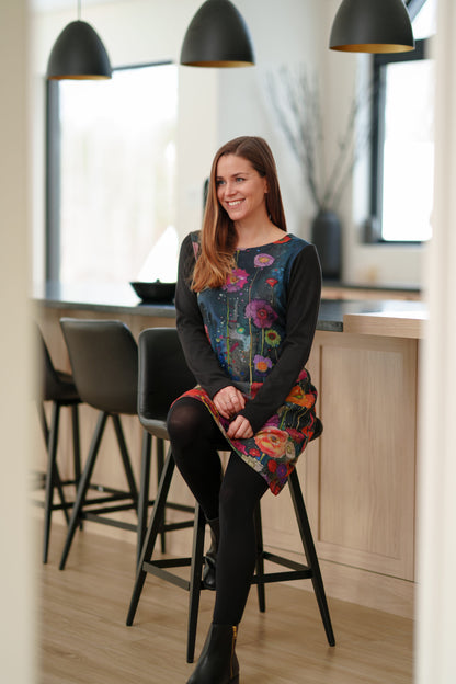 A woman wearing the Mistassini Dress by Rien ne se Perd in Floral Watercolour, an A-line knee-length dress with a vibrant floral print on the main body and contrasting black sleeves. She sitting by a counter in a kitchen setting. 