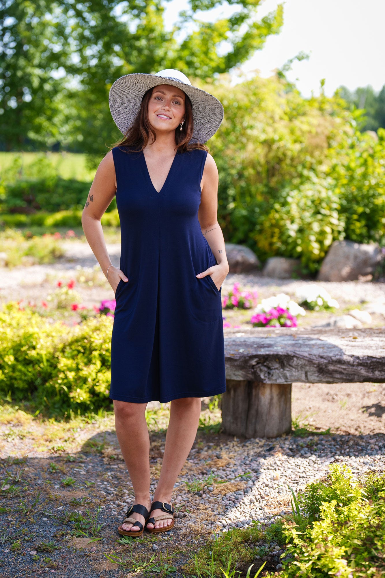 A woman wearing the Samba Dress by Rien ne se Perd in Navy, a sleeveless short dress with a V-neck, centre pleat, and side pockets. She is standing outside in a garden. 