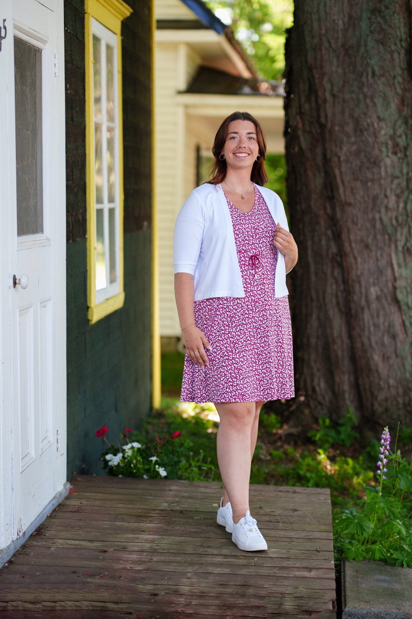 A woman wearing the Sangria Dress by Rien ne se Perd in Raspberry floral print, a short dress with a V-neck, an adjustable drawstring ribbon under the bust, and short sleeves. She is wearing it with a white bolero and standing on a door step with a tree behind her. 