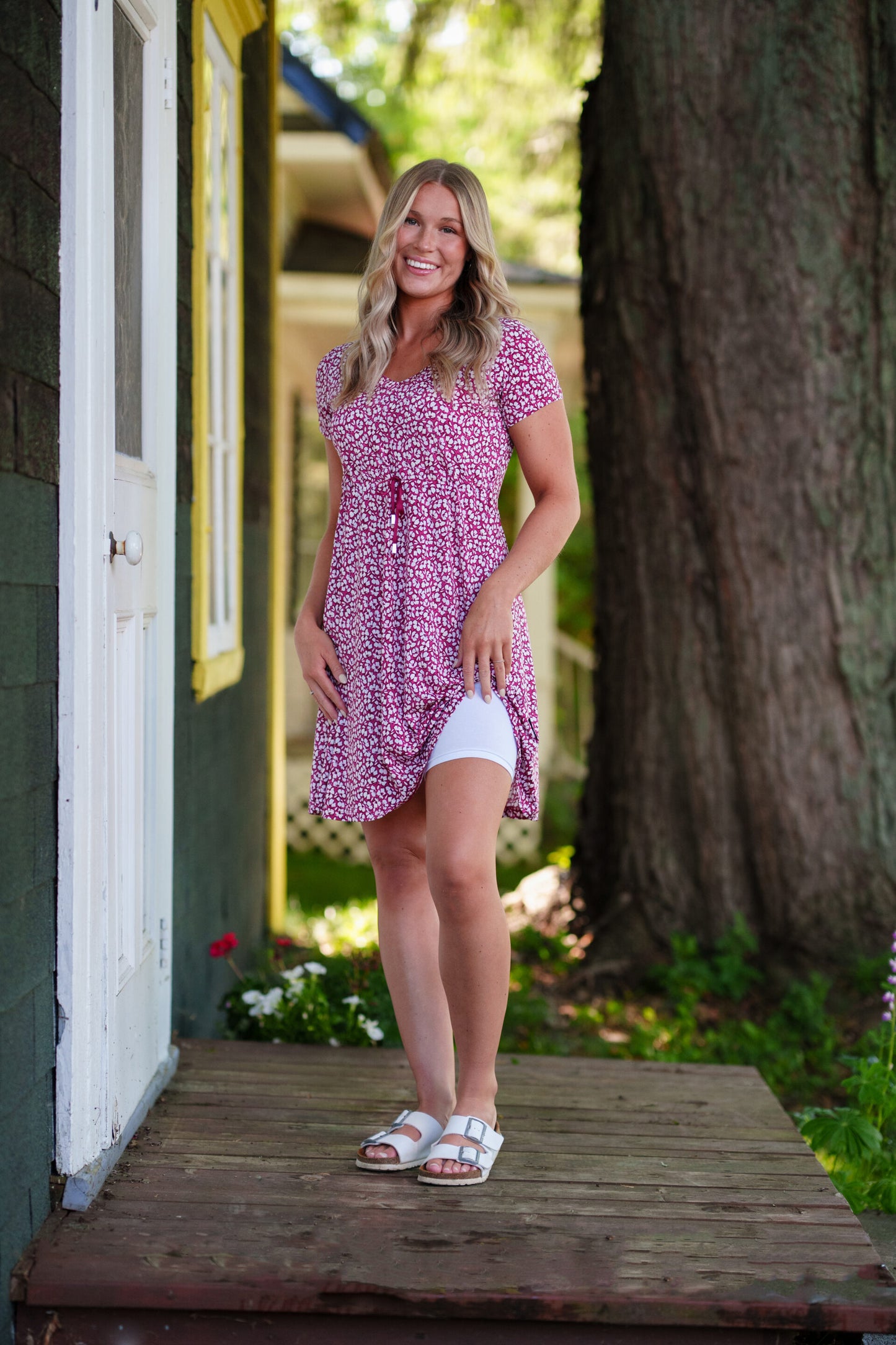 A woman wearing the Sangria Dress by Rien ne se Perd in Raspberry floral print, a short dress with a V-neck, an adjustable drawstring ribbon under the bust, and short sleeves. She is wearing it with white bike shorts underneath it and standing on a door step with a tree behind her. 