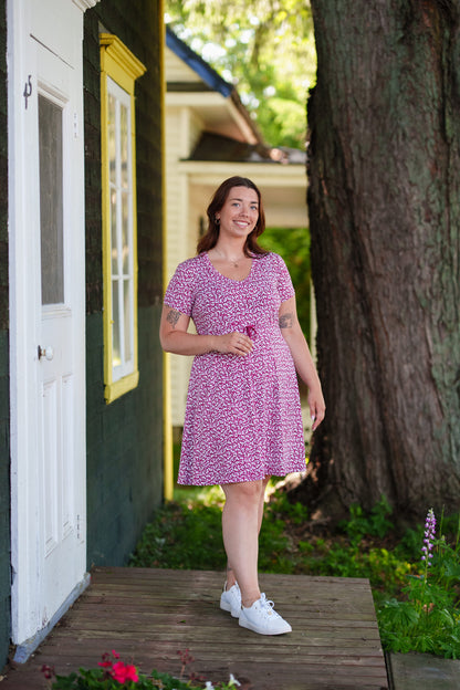 A woman wearing the Sangria Dress by Rien ne se Perd in Raspberry floral print, a short dress with a V-neck, an adjustable drawstring ribbon under the bust, and short sleeves. She is standing on a door step with a tree behind her. 