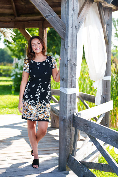 A woman wearing the Classic Dress by Rien ne se Perd in Black and Gold, a semi-fitted black  sheath dress with a gold floral pattern, short sleeves, and an above the knee length. She is standing outside in a gazebo.
