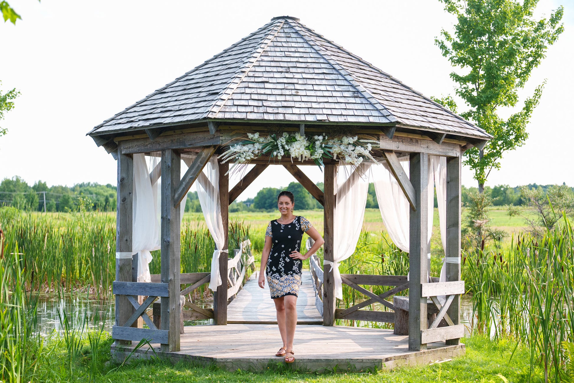 A woman wearing the Classic Dress by Rien ne se Perd in Black and Gold, a semi-fitted black  sheath dress with a gold floral pattern, short sleeves, and an above the knee length. She is standing outside in front of a wooden gazebo.
