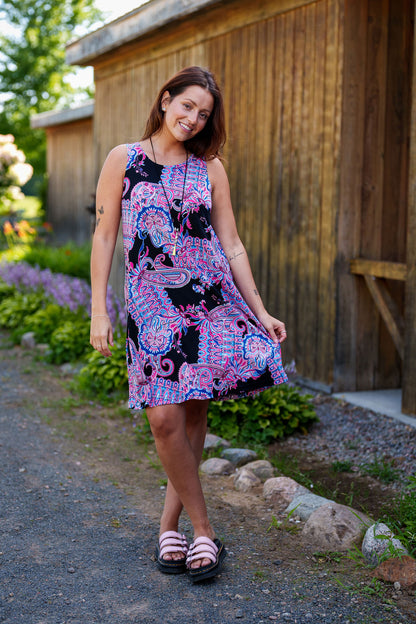 A woman wearing the Musette Dress by Rien ne se Perd, a short sleeveless swing dress with a purple Paisley print. She is standing in front of a wooden building. 