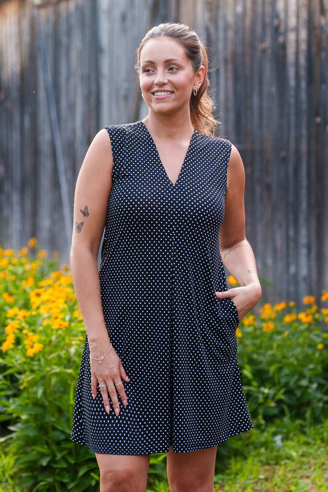 A 3/4 view of a woman wearing the Samba Dress by Rien ne se Perd in Black and White Polka Dot, a sleeveless short dress with a V-neck, centre pleat, and side pockets. She is standing outside in front of a wooden building. 