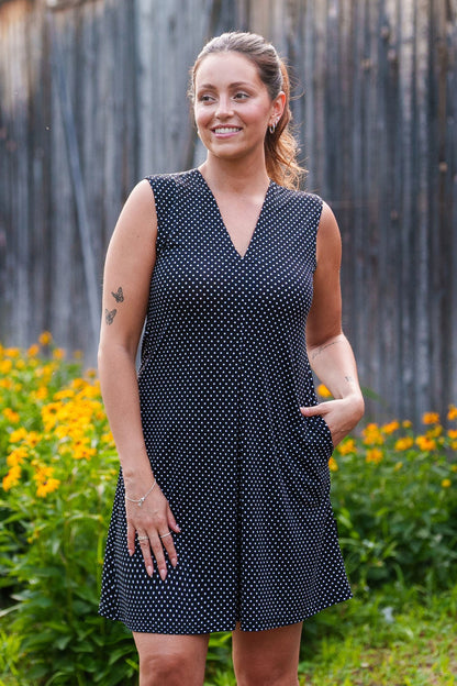 A 3/4 view of a woman wearing the Samba Dress by Rien ne se Perd in Black and White Polka Dot, a sleeveless short dress with a V-neck, centre pleat, and side pockets. She is standing outside in front of a wooden building. 