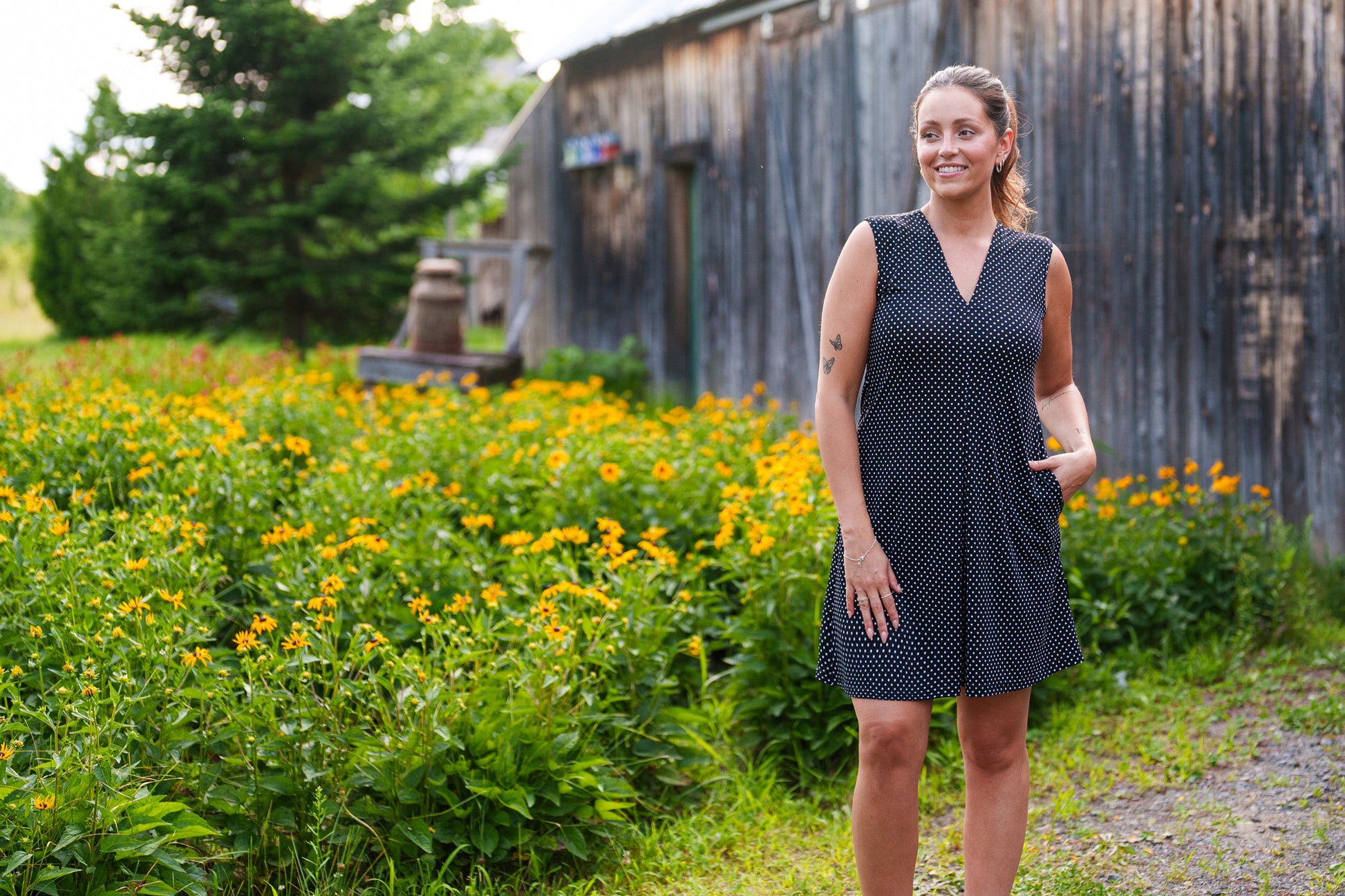 A 3/4 view of a woman wearing the Samba Dress by Rien ne se Perd in Black and White Polka Dots, a sleeveless short dress with a V-neck, centre pleat, and side pockets. She is standing outside in a garden. 
