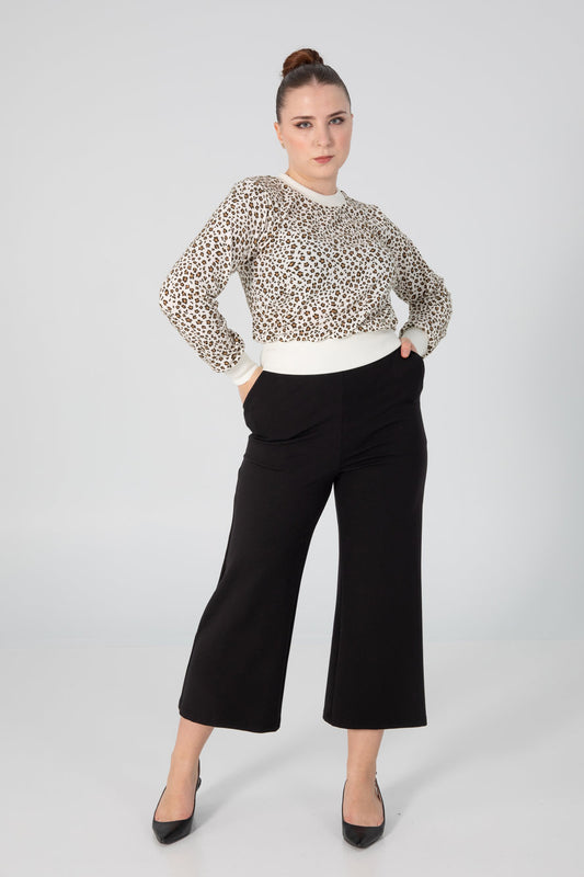 A woman wearing the Wanda Sweater by Slak in Leopard and Cream, featuring long sleeves, a round neck, a semi-fitted shape, and contrasting hem, cuffs and collar. She is wearing it with black pants and standing in front of a grey background. 