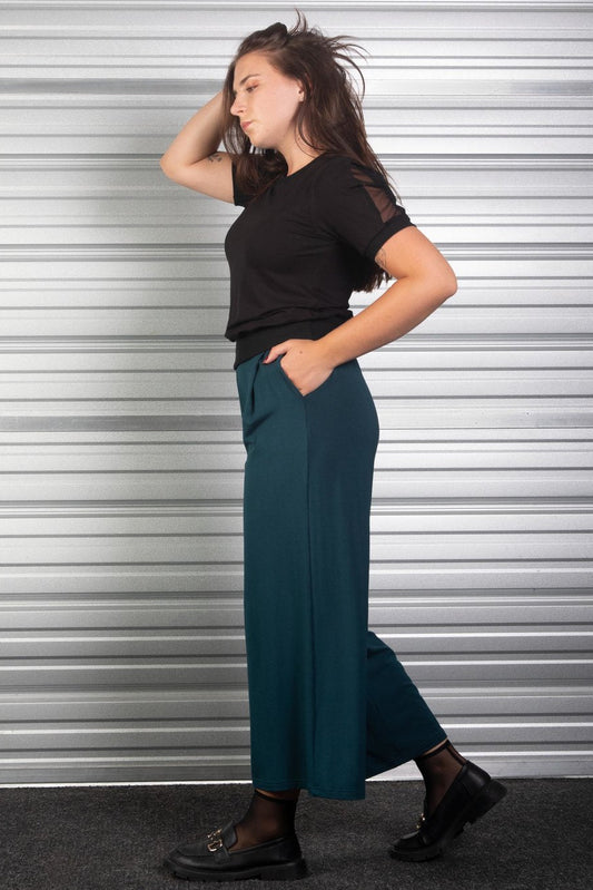Side view of a woman wearing the Darling Top by Slak in Black with teal pants, standing against a metallic shutter background