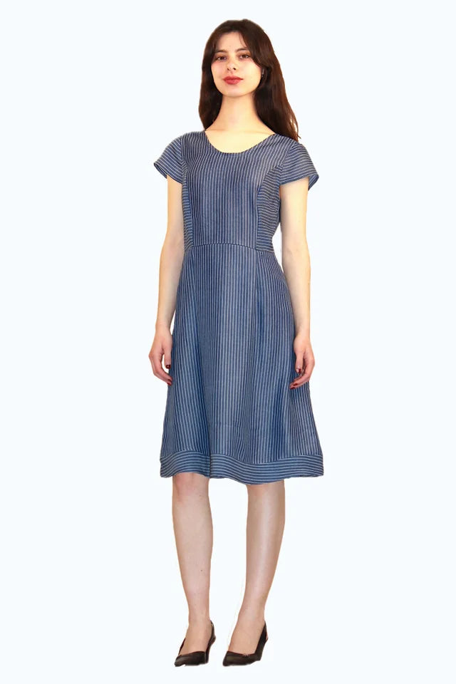 A woman wearing the Sequoia Dress by Ramonalisa in Striped denim, with a round neck, cap sleeves, a princess bodice, and an a-line knee-length skirt. She's standing in front of a white background. 