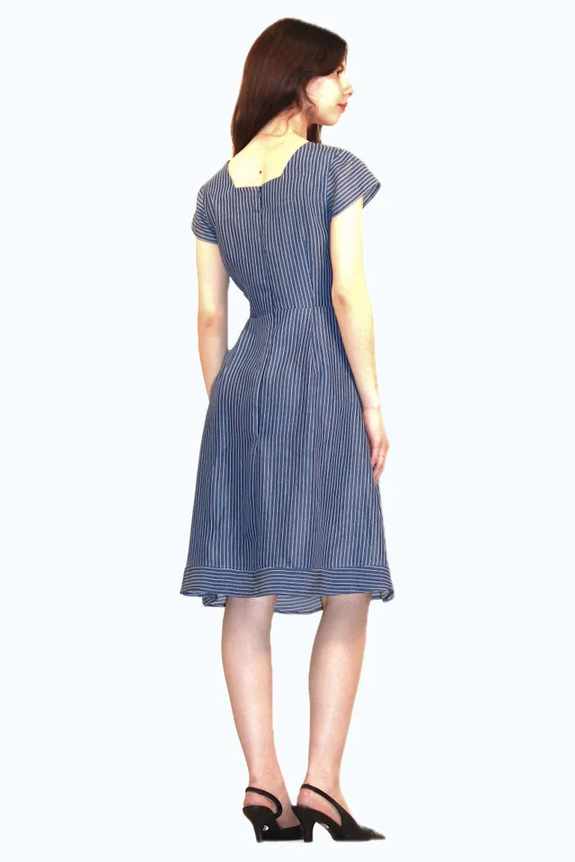 Back view of a woman wearing the Sequoia Dress by Ramonalisa in Striped denim, with a round neck, cap sleeves, a princess bodice, and an a-line knee-length skirt. She's standing in front of a white background. 