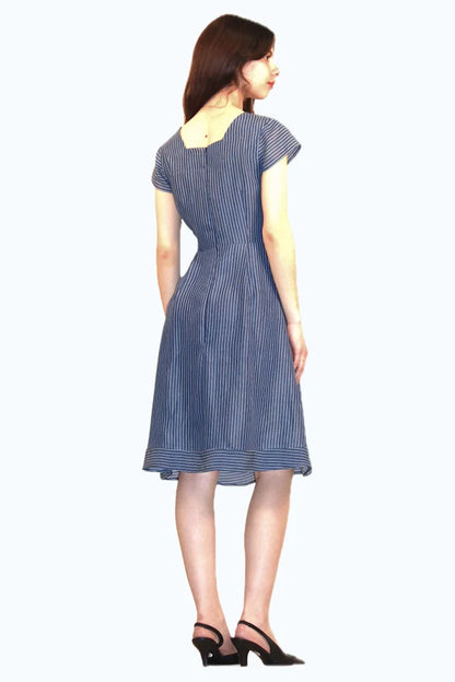 Back view of a woman wearing the Sequoia Dress by Ramonalisa in Striped denim, with a round neck, cap sleeves, a princess bodice, and an a-line knee-length skirt. She's standing in front of a white background. 