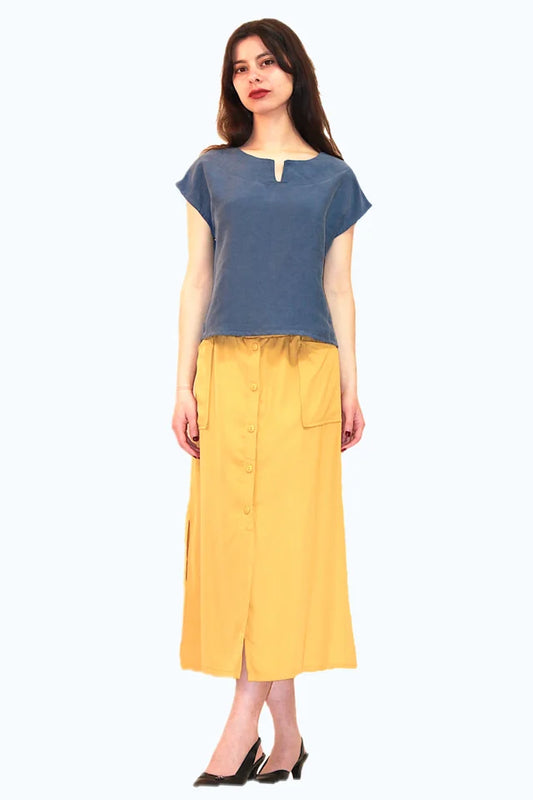 A woman wearing the Cassia Top by Ramonalisa in Cobalt, a short top with short drop-shoulder sleeves, and a round neck with a v-notch at the front. She is wearing it with a yellow skirt and standing in front of a white background. 