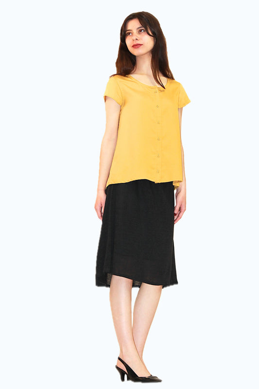 A woman wearing the Baobab Skirt by Ramonalisa in Black, featuring an elastic waist, below-the-knee hem that's curved at at the back, and a lining that's a little shorter than the main skirt. She is wearing it with the Juniper top in Mustard and standing in front of a white background. 