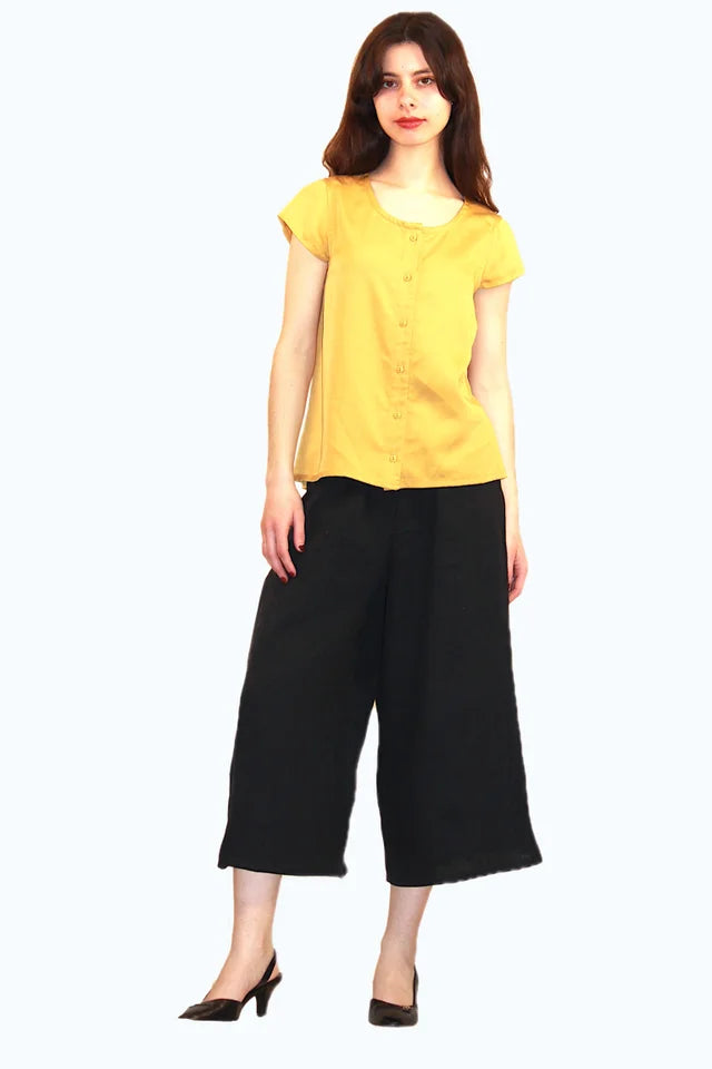 A woman wearing the Pagoda Pant by Ramonalisa, cropped, wide-legged linen pants with an elastic waist and side pockets. She is wearing them with the Juniper Top in Mustard and is standing in front of a white background. 