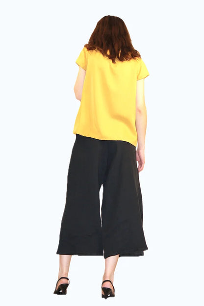 Back view of a woman wearing the Pagoda Pant by Ramonalisa, cropped, wide-legged linen pants with an elastic waist and side pockets. She is wearing them with the Juniper Top in Mustard and is standing in front of a white background. 