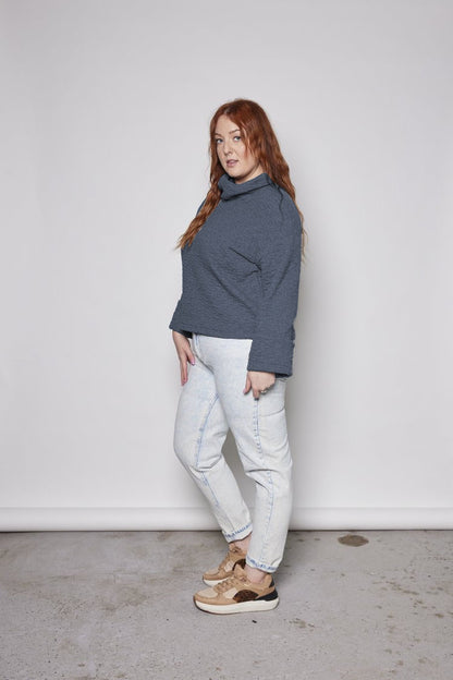 A woman wearing the Sable Top by Tangente in Dark Denim, a sweatshirt in quilted fabric with a cowl neck and cuffed sleeves and a slightly cropped and boxy fit. She is wearing it with jeans and standing in front of a white background. 