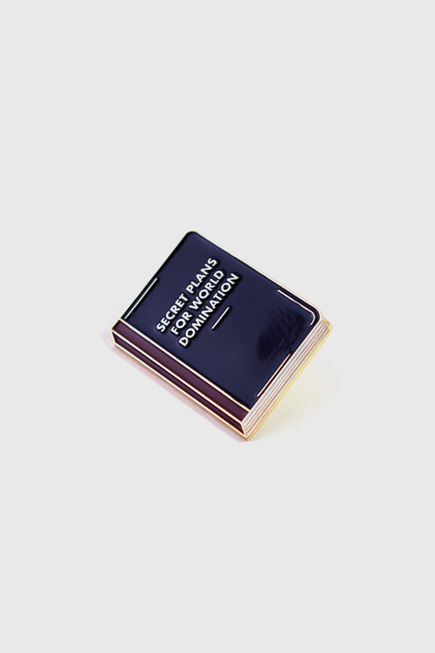 The World Domination Pin from Badaboöm Studio depicts a tiny book titled "Secret Plans for World Domination." It's shown against a white background. 