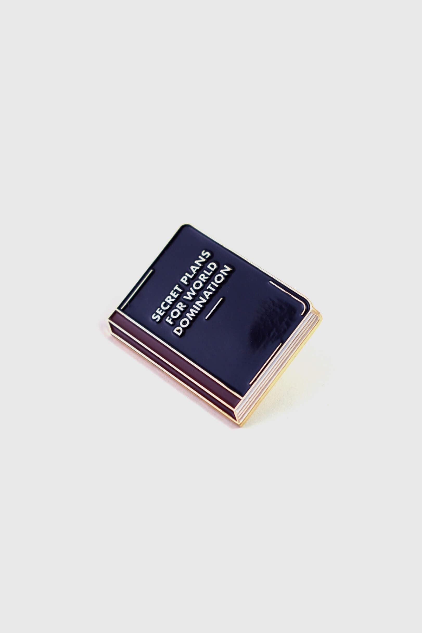 The World Domination Pin from Badaboöm Studio depicts a tiny book titled "Secret Plans for World Domination." It's shown against a white background. 