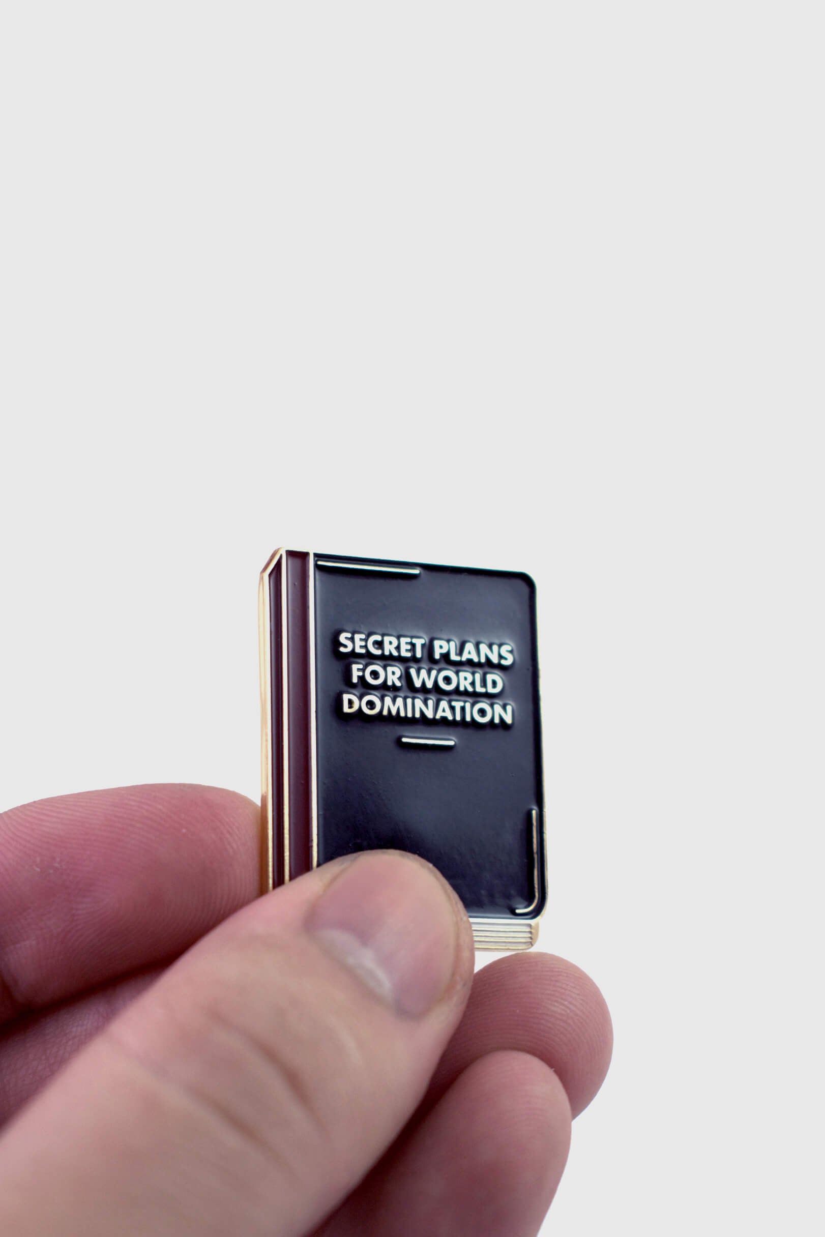A hand holding the World Domination Pin from Badaboöm Studio, a tiny book titled "Secret Plans for World Domination, is shown against a white background. 