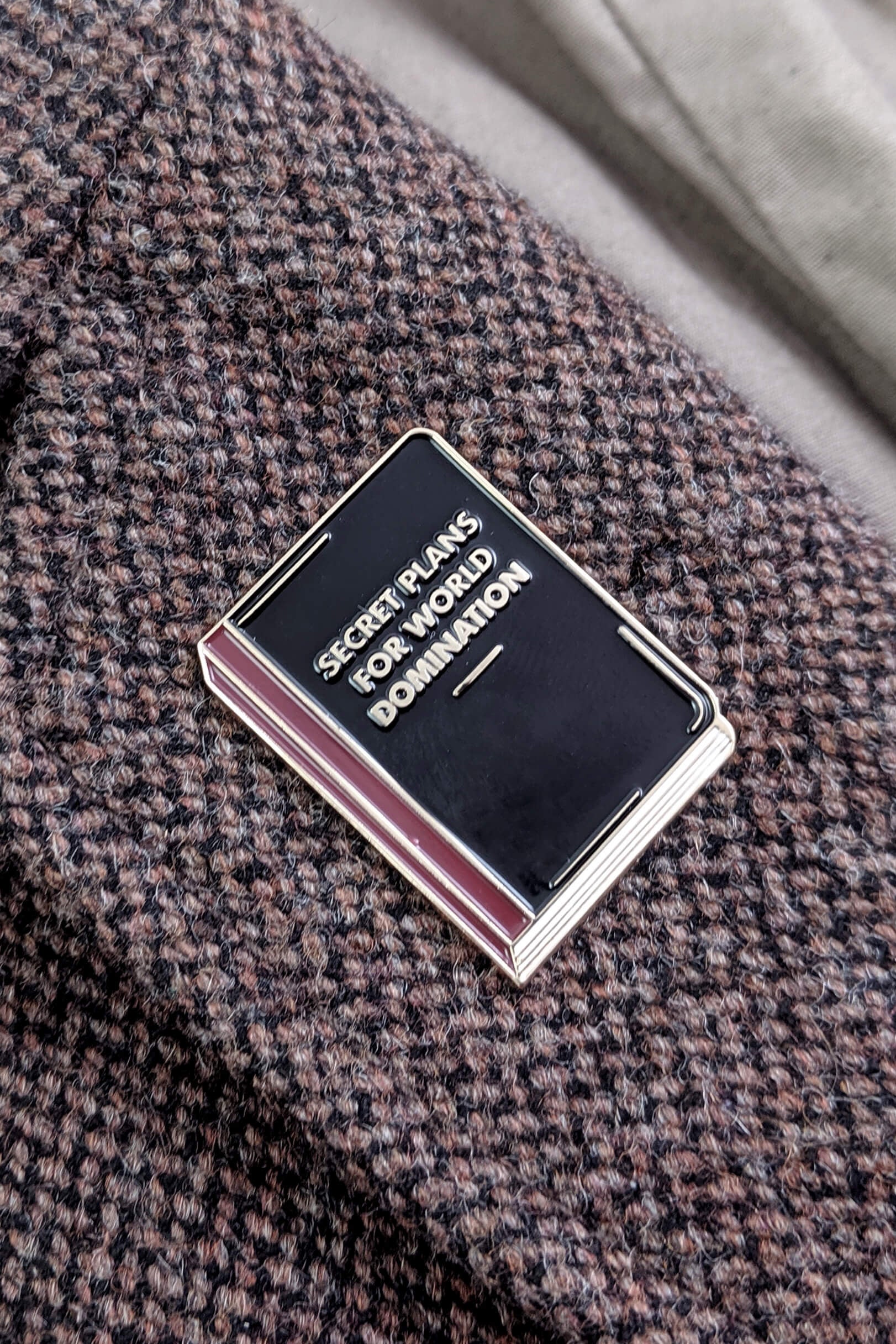 The World Domination Pin from Badaboöm Studio depicts a tiny book titled "Secret Plans for World Domination." It's shown against a tweed lapel.