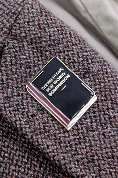The World Domination Pin from Badaboöm Studio depicts a tiny book titled "Secret Plans for World Domination." It's shown against a tweed lapel.