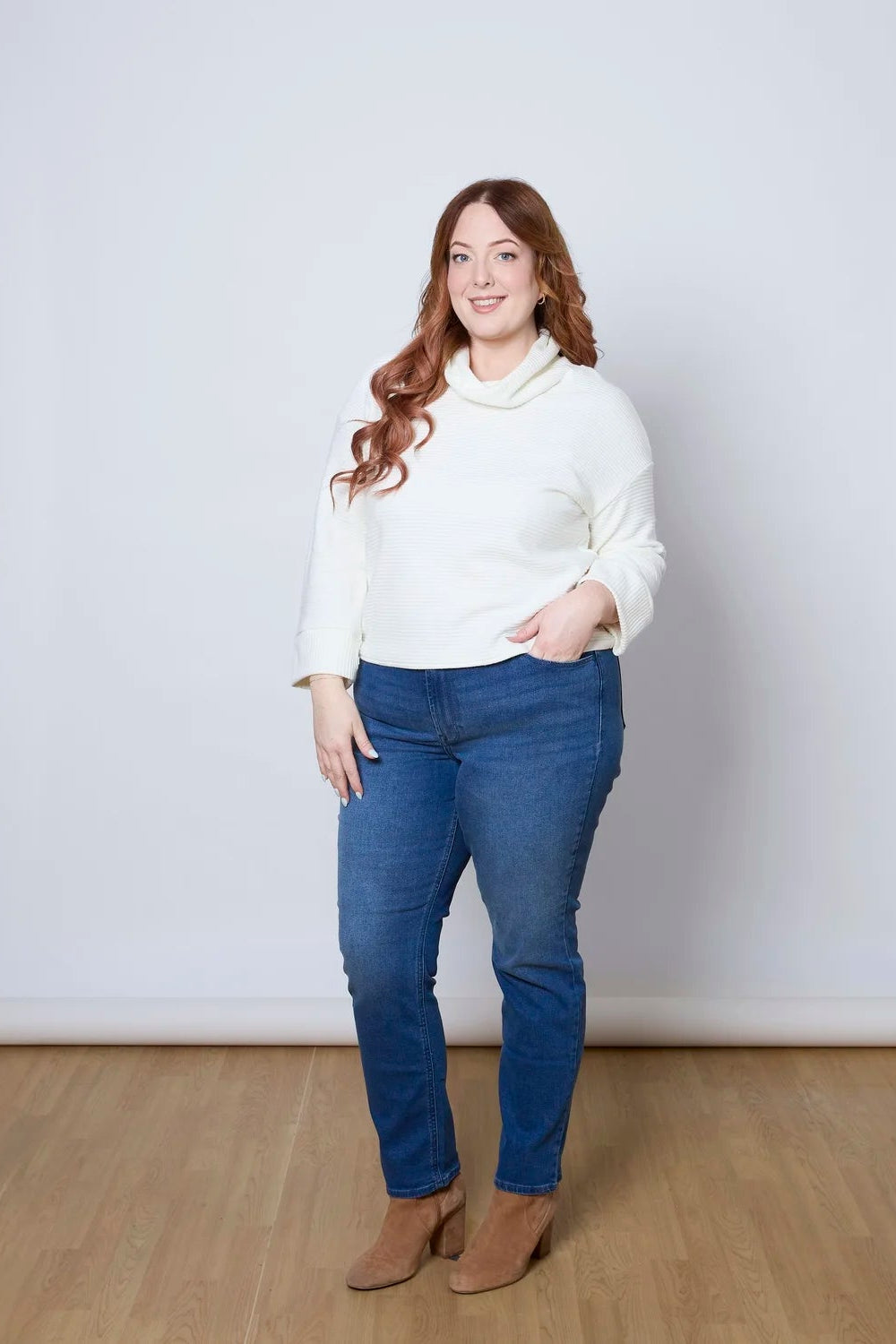 A woman wearing the Sidney Top by Tangente in Ivory, a slightly cropped, boxy top in ribbed fabric with long cuffed sleeves and a large cowl neck. She is wearing it with jeans and standing in front of a white wall. 