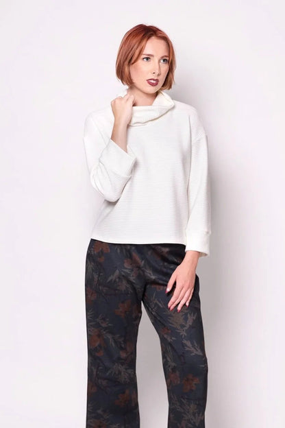 A woman wearing the Sidney Top by Tangente in Ivory, a slightly cropped, boxy top in ribbed fabric with long cuffed sleeves and a large cowl neck. She is wearing it with floral pants and standing in front of a white wall. 