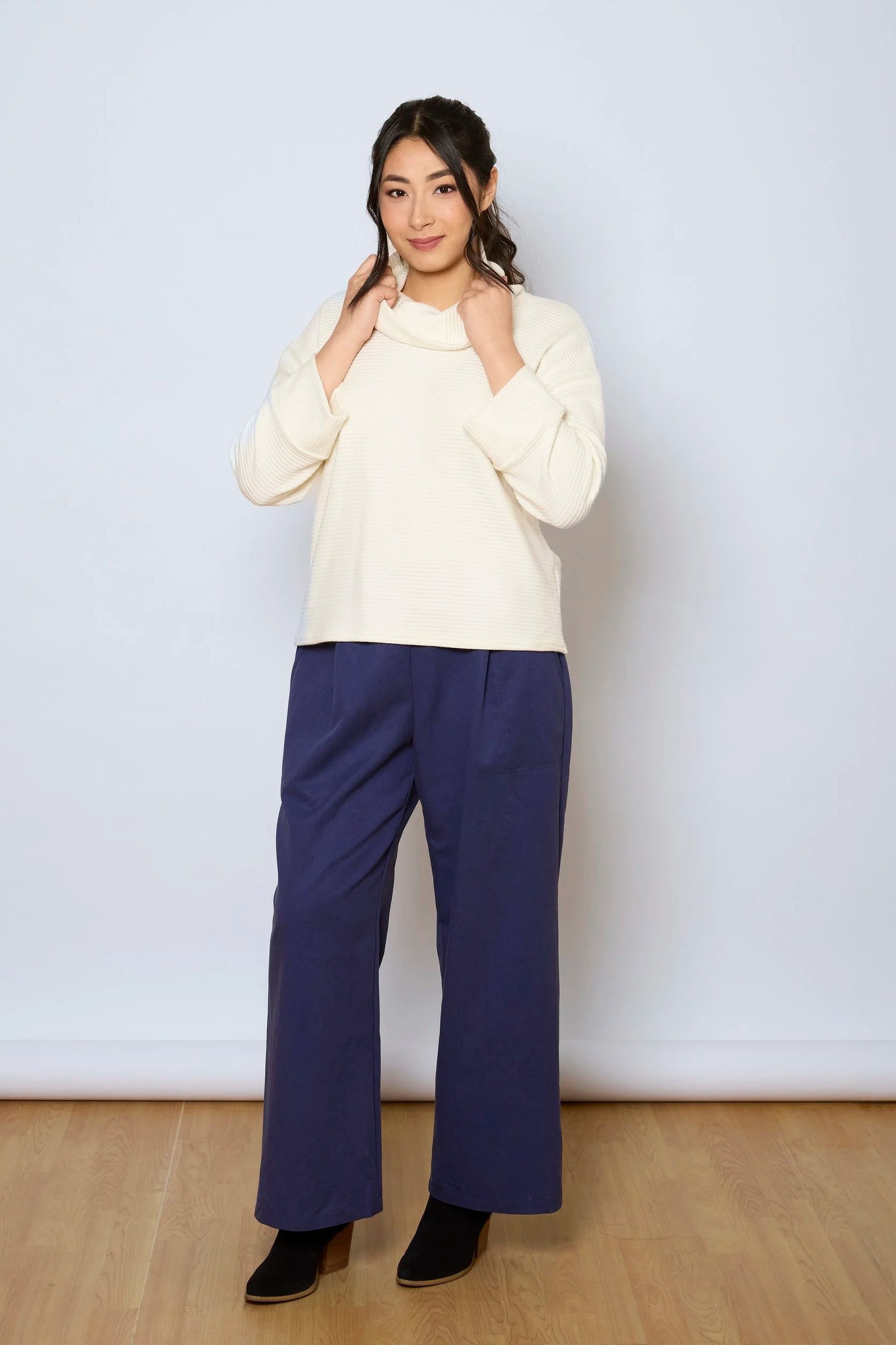 A woman wearing the Sidney Top by Tangente in Ivory, a slightly cropped, boxy top in ribbed fabric with long cuffed sleeves and a large cowl neck. She is wearing it with navy pants and standing in front of a white wall. 